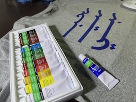 A set of acrylic paint tubes is arranged neatly in a white plastic tray, with various colors labeled on each tube. In the background, there is a grey fabric or t-shirt featuring blue calligraphy or script. A single tube of paint is placed beside the tray.