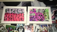 Two framed graffiti artworks display abstract and colorful designs. The left artwork features a vibrant pink background with black outlines creating curvy, overlapping shapes. The right artwork has a mix of purple and green tones with bold black outlines forming abstract faces and repetitive text. The frames rest on a surface covered with various stickers and cutouts, adding to the urban and eclectic aesthetic.