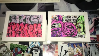 Two framed graffiti artworks display abstract and colorful designs. The left artwork features a vibrant pink background with black outlines creating curvy, overlapping shapes. The right artwork has a mix of purple and green tones with bold black outlines forming abstract faces and repetitive text. The frames rest on a surface covered with various stickers and cutouts, adding to the urban and eclectic aesthetic.