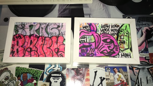 Two framed graffiti artworks display abstract and colorful designs. The left artwork features a vibrant pink background with black outlines creating curvy, overlapping shapes. The right artwork has a mix of purple and green tones with bold black outlines forming abstract faces and repetitive text. The frames rest on a surface covered with various stickers and cutouts, adding to the urban and eclectic aesthetic.