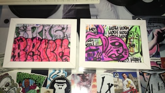 Two framed graffiti artworks display abstract and colorful designs. The left artwork features a vibrant pink background with black outlines creating curvy, overlapping shapes. The right artwork has a mix of purple and green tones with bold black outlines forming abstract faces and repetitive text. The frames rest on a surface covered with various stickers and cutouts, adding to the urban and eclectic aesthetic.