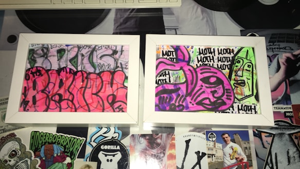 Two framed graffiti artworks display abstract and colorful designs. The left artwork features a vibrant pink background with black outlines creating curvy, overlapping shapes. The right artwork has a mix of purple and green tones with bold black outlines forming abstract faces and repetitive text. The frames rest on a surface covered with various stickers and cutouts, adding to the urban and eclectic aesthetic.