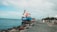A large cargo ship with the name 'Peter Ronna Gibraltar' is docked along a rocky shoreline. The ship is predominantly blue and white, featuring a visible safety boat attached to its side. The surrounding area includes a paved walkway lined with palm trees and a few orange traffic cones. The sky is overcast, and there are cranes and buildings in the distant background.