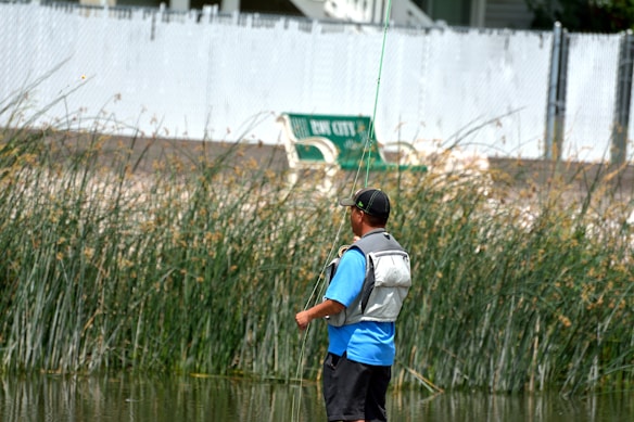 A person stands near a body of water, dressed in a blue shirt and a life vest, and appears to be fishing. Tall grass and reeds are visible in the background, along with a bench and a fence. The setting seems relaxed and outdoor-oriented.