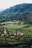 A lush green tea plantation stretches across rolling hills with a group of people gathered on a designated path. The landscape is marked by neatly arranged rows of tea plants. A sign indicates the name 'Boh Tea' on a hillside. The backdrop includes dense forests and misty hills, giving a serene and expansive view.