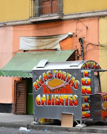 A small food stand is positioned against a colorful urban wall. The stand prominently displays brightly colored signage advertising 'Ricas Tortas Calientes.' Above the stand, there is a faded and torn awning, and a mix of green and orange hues paint the surrounding walls. The environment appears slightly messy with some discarded items on the pavement.