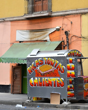A small food stand is positioned against a colorful urban wall. The stand prominently displays brightly colored signage advertising 'Ricas Tortas Calientes.' Above the stand, there is a faded and torn awning, and a mix of green and orange hues paint the surrounding walls. The environment appears slightly messy with some discarded items on the pavement.
