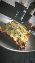 A plate of lasagna topped with melted cheese and herbs is placed on a dark woven placemat. In the background, there is a partially filled glass of red wine and a bottle of wine. A loaf of bread is also visible next to the wine bottle.