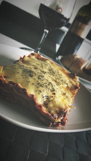 A plate of lasagna topped with melted cheese and herbs is placed on a dark woven placemat. In the background, there is a partially filled glass of red wine and a bottle of wine. A loaf of bread is also visible next to the wine bottle.