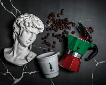 A white marble bust of a classical figure is placed next to a white cup with the word 'Italy' and a moka pot in the colors of the Italian flag. Coffee beans are scattered on a dark marble surface around these objects.
