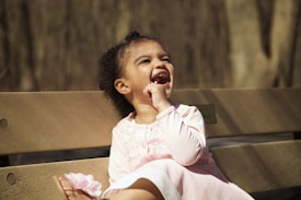 A young child sits on a wooden bench outdoors, laughing joyfully. The child is wearing a light pink dress and has curly dark hair. The background shows blurred trees, suggesting a park setting.
