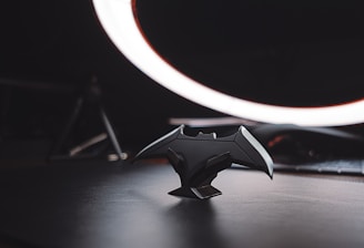 A dark themed setting features a bat-shaped figurine placed on a surface in the foreground, illuminated dramatically by a circular light source in the background. The figurine is detailed, capturing the silhouettes of bat wings and other angular elements. The environment appears minimalistic with an emphasis on the central object, while the background remains blurred, highlighting the focus on the bat shape.