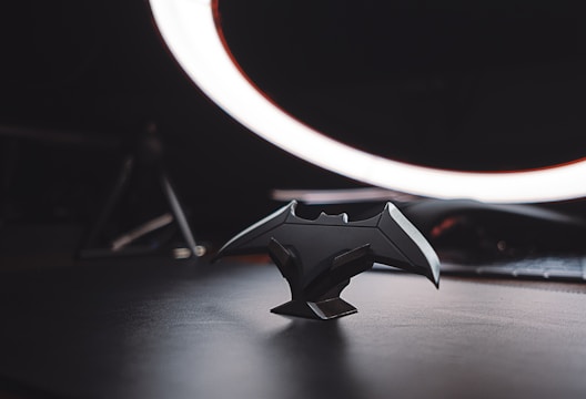 A dark themed setting features a bat-shaped figurine placed on a surface in the foreground, illuminated dramatically by a circular light source in the background. The figurine is detailed, capturing the silhouettes of bat wings and other angular elements. The environment appears minimalistic with an emphasis on the central object, while the background remains blurred, highlighting the focus on the bat shape.