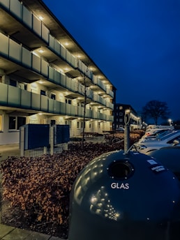 A nighttime scene featuring a modern apartment building with illuminated balconies and a row of parked cars. A large spherical recycling container labeled 'GLAS' is in the foreground, surrounded by bare trees and hedges. The sky is a deep blue, providing a contrasting background to the well-lit structure.