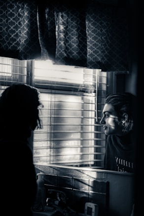 A person is looking at their reflection in a mirror near a window with blinds partially open, allowing in some light. The image is in black and white, creating a moody and introspective atmosphere. On the counter, various items such as a bottle and a small device are visible. Curtains with a patterned design are partly hanging above the window.