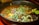 A vibrant, appetizing bowl of soup garnished with fresh greens, sprouts, and various colorful vegetables including sliced cucumber and pieces of bright yellow bell peppers. The broth is rich and dark, surrounded by a black bowl. Black sesame seeds are sprinkled across the surface, adding texture and contrast.