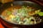 A vibrant, appetizing bowl of soup garnished with fresh greens, sprouts, and various colorful vegetables including sliced cucumber and pieces of bright yellow bell peppers. The broth is rich and dark, surrounded by a black bowl. Black sesame seeds are sprinkled across the surface, adding texture and contrast.