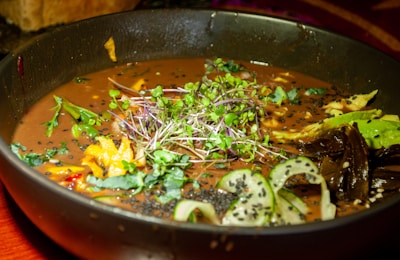 A vibrant, appetizing bowl of soup garnished with fresh greens, sprouts, and various colorful vegetables including sliced cucumber and pieces of bright yellow bell peppers. The broth is rich and dark, surrounded by a black bowl. Black sesame seeds are sprinkled across the surface, adding texture and contrast.
