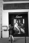 A black and white photograph of a storefront window featuring a large poster for Etam. The poster depicts a woman lounging in lingerie. A woman is walking past on the sidewalk, wearing a patterned dress and high heels.