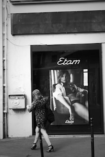 A black and white photograph of a storefront window featuring a large poster for Etam. The poster depicts a woman lounging in lingerie. A woman is walking past on the sidewalk, wearing a patterned dress and high heels.