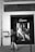 A black and white photograph of a storefront window featuring a large poster for Etam. The poster depicts a woman lounging in lingerie. A woman is walking past on the sidewalk, wearing a patterned dress and high heels.