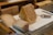 A variety of cheeses is arranged on a white cutting board. The cheeses have different colors and textures, with some appearing aged and others speckled with herbs. A cheese slicer is placed beside them on the board, with a wooden and stainless steel table surface underneath.