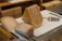 A variety of cheeses is arranged on a white cutting board. The cheeses have different colors and textures, with some appearing aged and others speckled with herbs. A cheese slicer is placed beside them on the board, with a wooden and stainless steel table surface underneath.