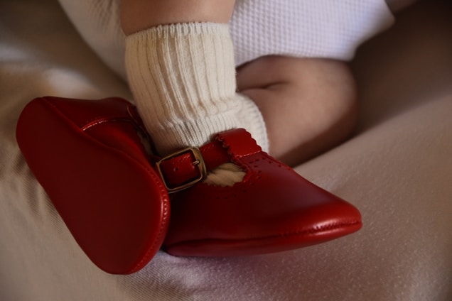 A baby's foot is wearing a bright red shoe with a buckle, accompanied by a white sock. The baby is dressed in a textured, cream-colored garment, and their skin appears soft and smooth.