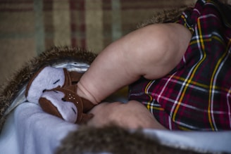 A close-up of a baby's legs dressed in a plaid-patterned outfit. The baby is wearing soft animal-themed slippers with a furry lining visible. The background has a textured pattern in muted colors.