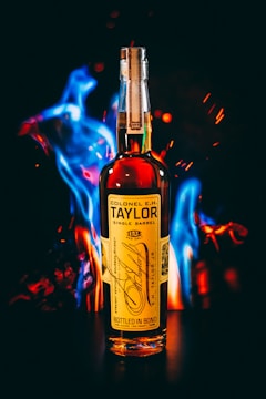 A bottle of Colonel E.H. Taylor Single Barrel bourbon is positioned against a dark background with vibrant blue and orange flames surrounding it. The label is detailed, featuring gold and black text with a signature prominently displayed. The flames create a dramatic contrast, highlighting the bottle.