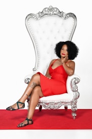 A woman wearing a bright red dress sits confidently on an ornate silver and white throne chair. Her expression is poised, with one hand resting on her face. The setting includes a red carpet under the chair, enhancing the overall luxurious appearance.