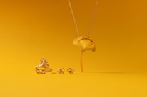 A collection of gold jewelry is set against a vibrant yellow background. The pieces include a ring, earrings, and a necklace, all designed to resemble a ginkgo leaf. The shiny metallic finish of the jewelry contrasts with the matte yellow surface.