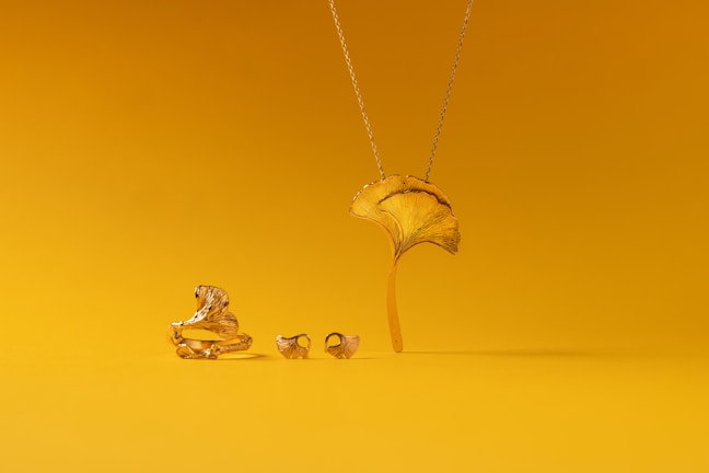 A collection of gold jewelry is set against a vibrant yellow background. The pieces include a ring, earrings, and a necklace, all designed to resemble a ginkgo leaf. The shiny metallic finish of the jewelry contrasts with the matte yellow surface.