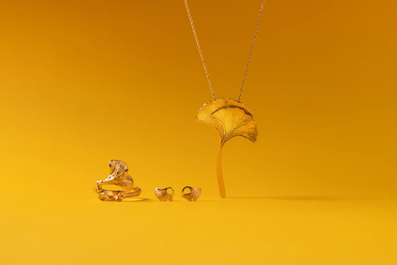 A collection of gold jewelry is set against a vibrant yellow background. The pieces include a ring, earrings, and a necklace, all designed to resemble a ginkgo leaf. The shiny metallic finish of the jewelry contrasts with the matte yellow surface.