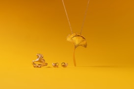 A collection of gold jewelry is set against a vibrant yellow background. The pieces include a ring, earrings, and a necklace, all designed to resemble a ginkgo leaf. The shiny metallic finish of the jewelry contrasts with the matte yellow surface.