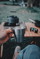A person holding a stainless steel mug while sitting outdoors. A portable gas stove with a kettle is on a wooden table nearby, and a camera is visible in the background. The scene captures a relaxed outdoor setting.