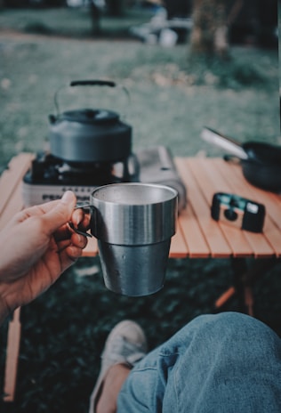 A person holding a stainless steel mug while sitting outdoors. A portable gas stove with a kettle is on a wooden table nearby, and a camera is visible in the background. The scene captures a relaxed outdoor setting.