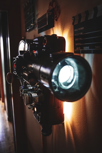 A vintage film projector is mounted on a wall, casting light into the room. The mechanical details of the projector are visible, including reels, dials, and knobs. Nearby, a clapperboard with filmmaking details is attached to the wall. The lighting is warm, highlighting the retro and cinematic atmosphere.