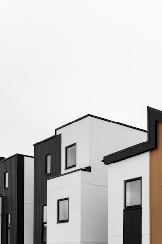 Minimalist architecture featuring modern buildings with flat roofs and clean lines. The structures are predominantly white with black and wooden panel accents. Large, square windows punctuate the facades.