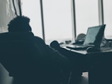 A person is reclining in a chair with their back to the camera, looking at a laptop on a desk. The setting appears to be an office or workspace with large windows providing natural light. Papers and various desk items are scattered around, creating a casual, relaxed environment.
