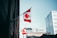 A Canadian flag is prominently displayed on a flagpole affixed to a tall brick building. The flag features a red maple leaf on a white background flanked by two red bands. In the background, modern office buildings with glass and concrete structures are visible under a clear blue sky.
