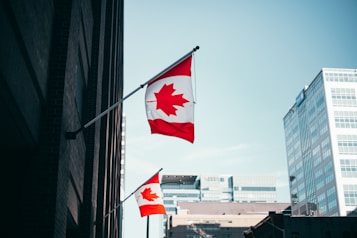 A Canadian flag is prominently displayed on a flagpole affixed to a tall brick building. The flag features a red maple leaf on a white background flanked by two red bands. In the background, modern office buildings with glass and concrete structures are visible under a clear blue sky.