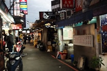 A busy street market scene with several food stalls displaying menus in Chinese script. Signs for various brands like Reebok, Nike, and Adidas are visible. People are walking along the narrow aisle, and scooters are parked along the sides.