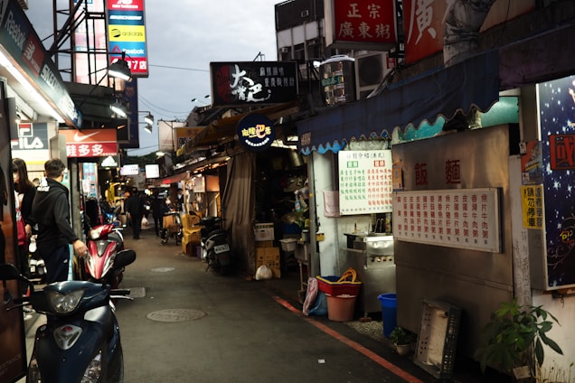 A busy street market scene with several food stalls displaying menus in Chinese script. Signs for various brands like Reebok, Nike, and Adidas are visible. People are walking along the narrow aisle, and scooters are parked along the sides.