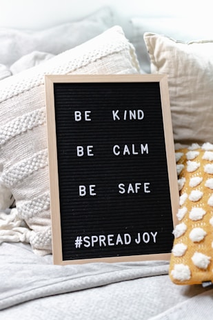 A cozy setting featuring a letter board with a wooden frame, displaying inspirational messages: 'BE KIND', 'BE CALM', 'BE SAFE', and '#SPREADJOY'. It is placed on a soft, textured surface alongside textured pillows, including one with a yellow and white polka dot pattern.