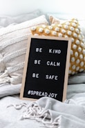 A black letter board with a wooden frame displays the words 'BE KIND, BE CALM, BE SAFE #SPREADJOY'. It is propped up on a cozy arrangement of textured cushions and a soft, fringed blanket in neutral colors.