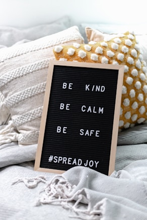 A black letter board with a wooden frame displays the words 'BE KIND, BE CALM, BE SAFE #SPREADJOY'. It is propped up on a cozy arrangement of textured cushions and a soft, fringed blanket in neutral colors.