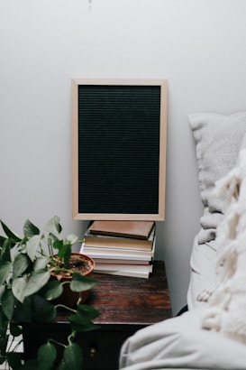 A cozy interior scene featuring a wooden table with a small stack of books topped by a blank chalkboard. A potted plant with green leaves sits adjacent to the books, adding a touch of nature. A soft white pillow or blanket is partially visible on a bed or couch to the right.