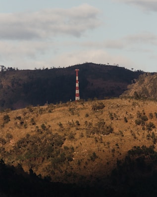 A red and white communications tower stands on the slope of a hilly landscape covered with sparse vegetation. The foreground features rolling hills with scattered trees, while a darker forested area extends along the background. Overhead, the sky is partly cloudy with soft lighting casting shadows on the terrain.