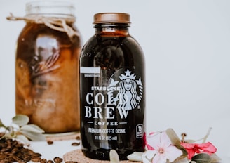 A bottle of Starbucks Cold Brew coffee is prominently displayed alongside a jar filled with coffee beans. Decorative flowers and leaves adorn the scene, adding elegance. Coffee beans are scattered around for an aesthetic touch.